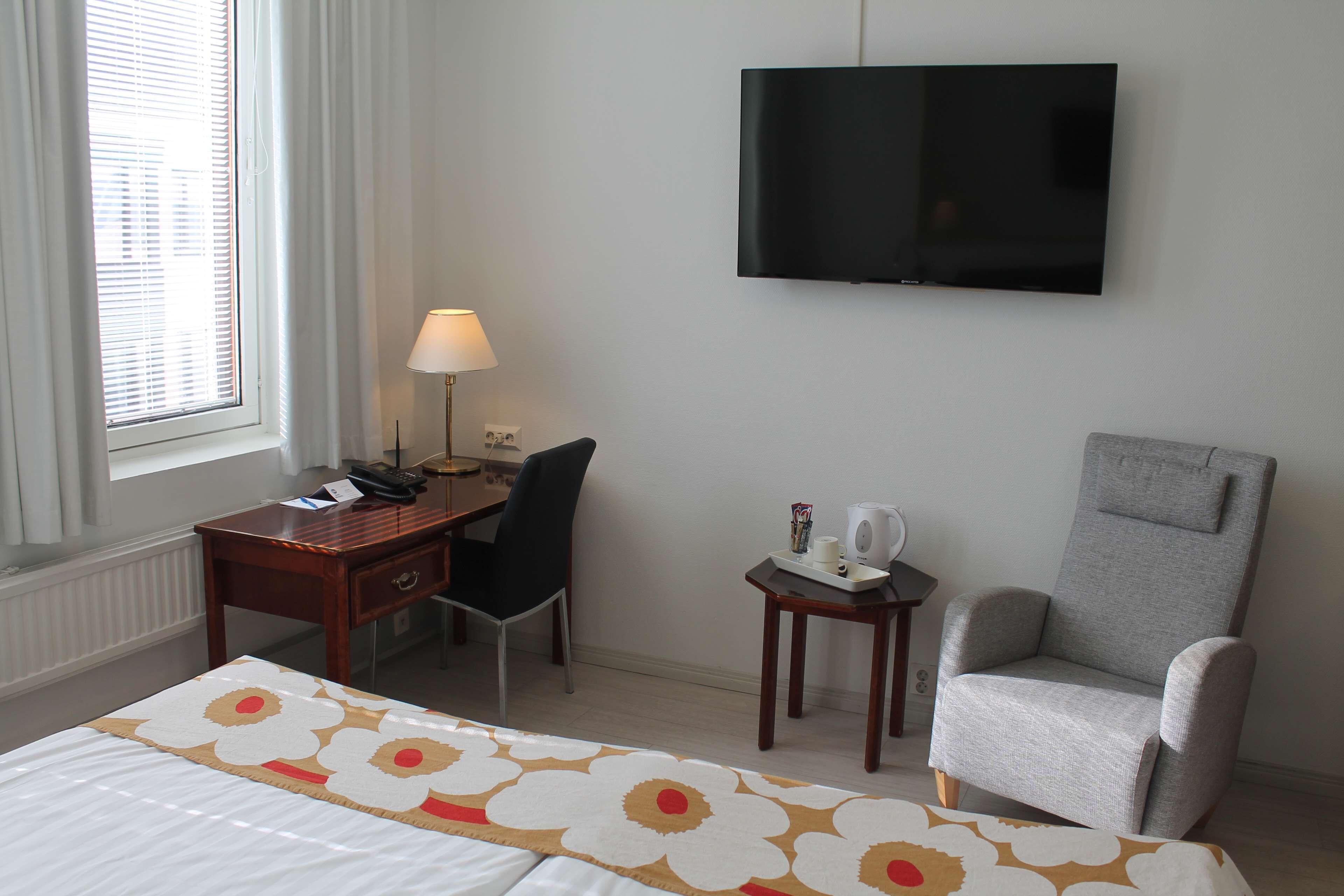 Habitación de hotel con cama, escritorio con silla, televisor y una mesita de noche con lámpara.