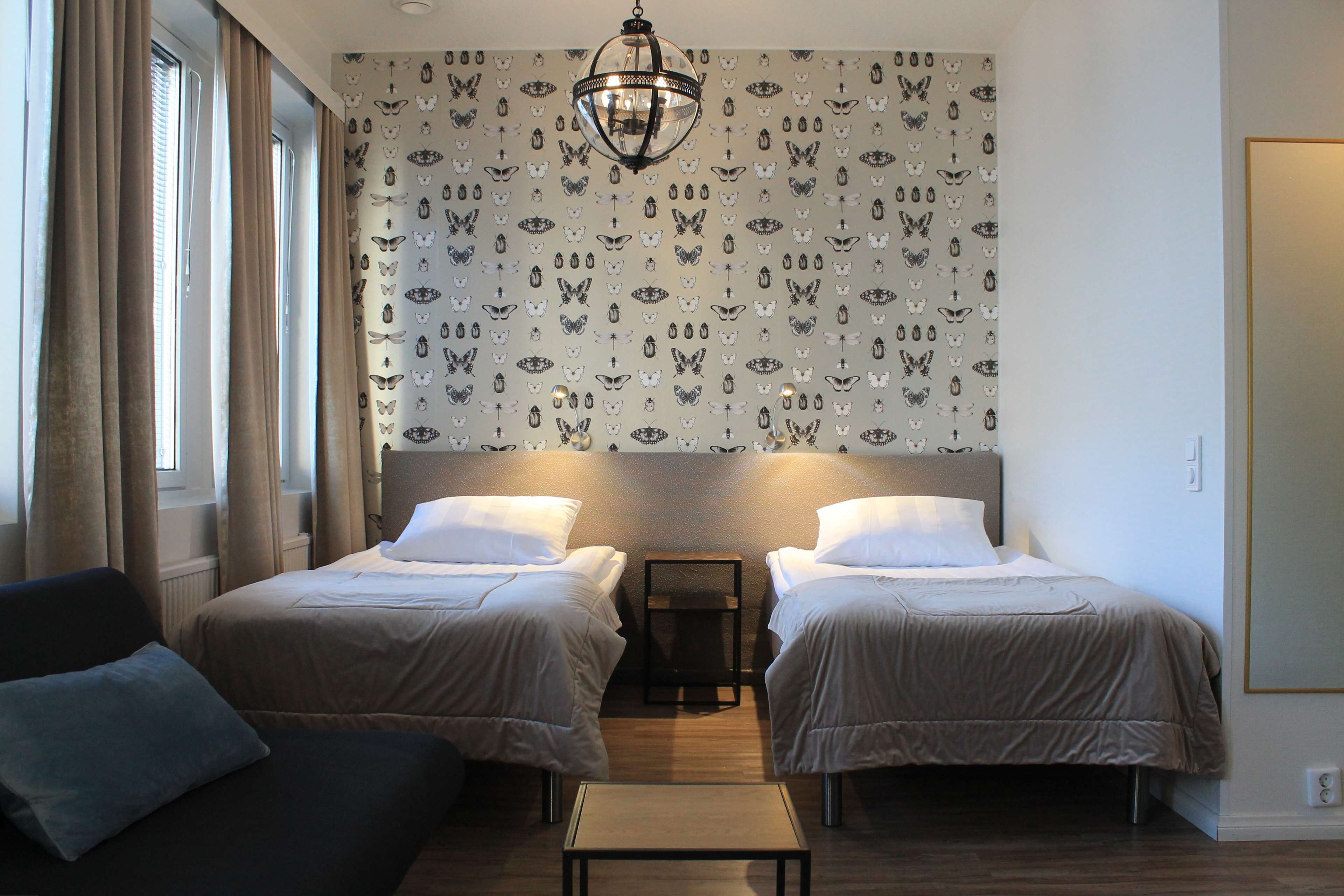 Ein Hotelzimmer mit zwei Betten, einem kleinen Tisch und einer Couch. Die Wände sind mit einem gemusterten Papier tapeziert und ein Chandelier hängt von der Decke.