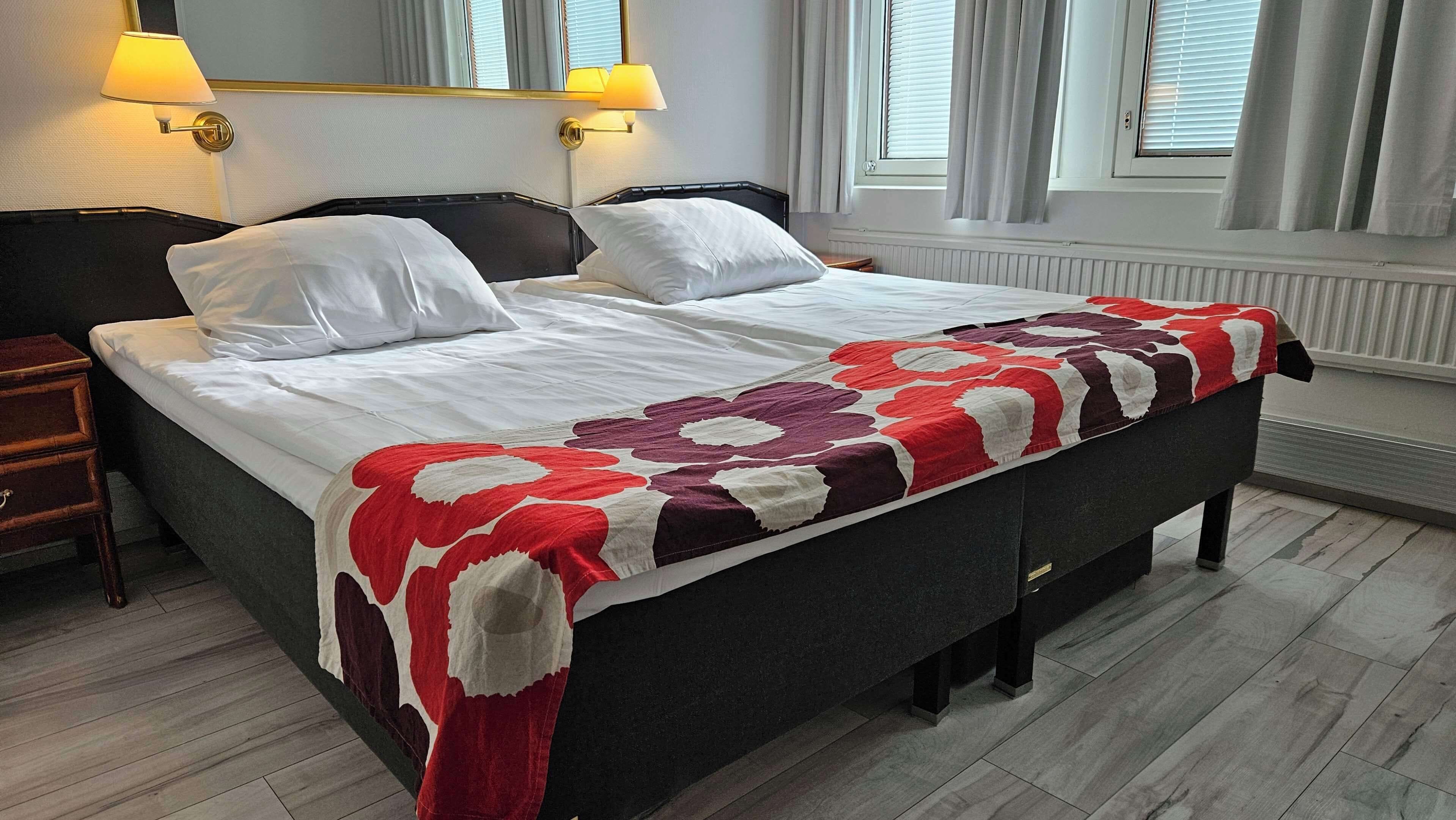Una cama doble con una colcha estampada, dos lámparas de noche, una mesita de noche y un espejo grande sobre la cama.