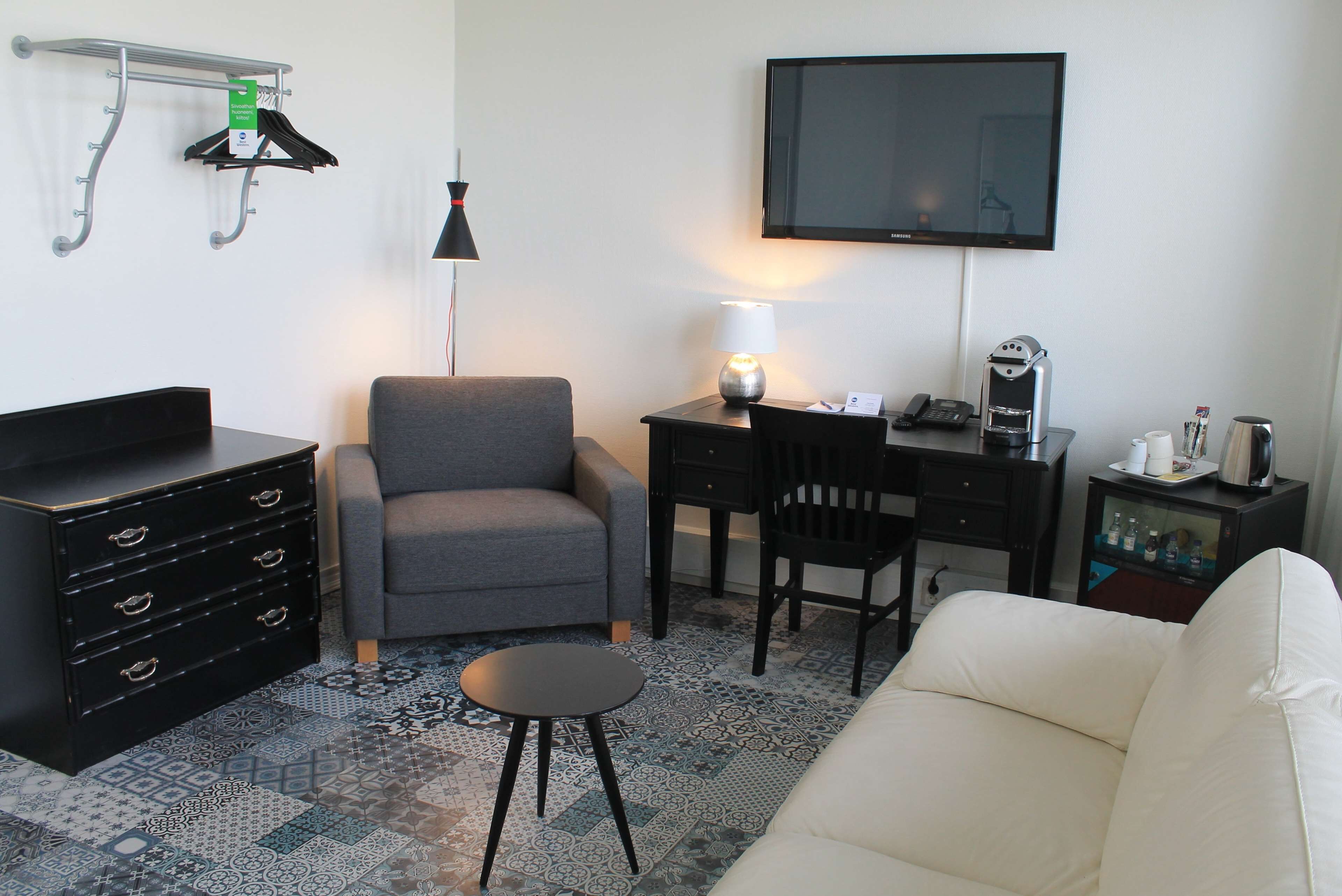 Un espace de vie compact comprenant un fauteuil gris, une petite table ronde, un canapé blanc, un bureau avec une chaise, un téléviseur à écran plat, une lampe, une machine à café et un meuble de rangement avec des vêtements suspendus.