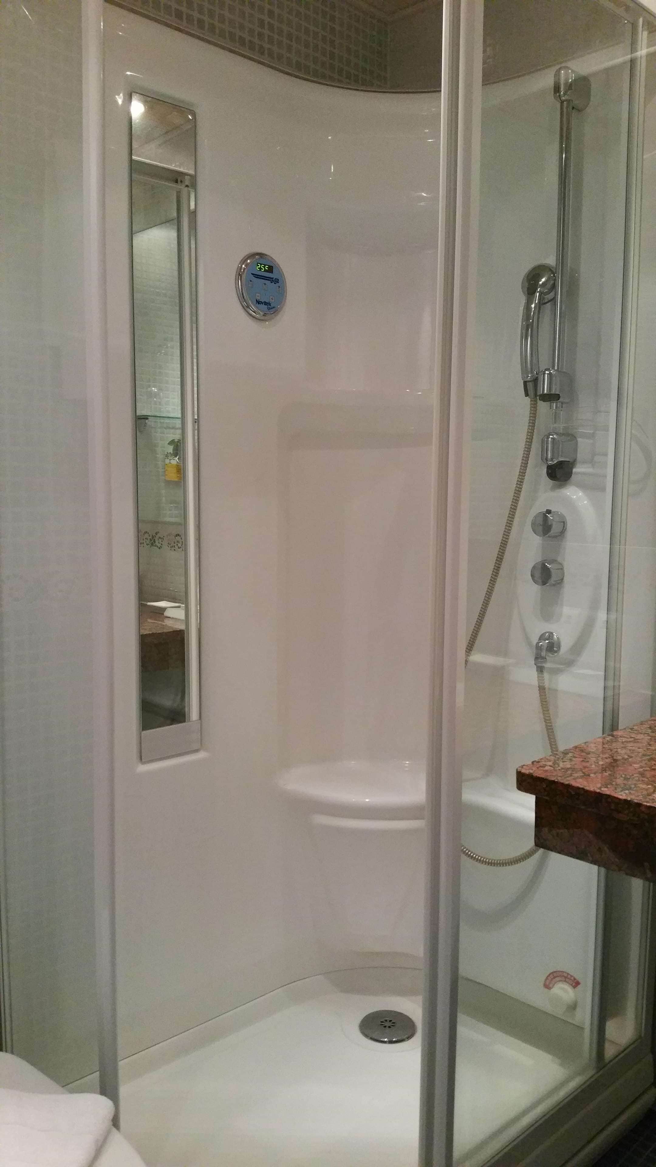 Cabina de ducha con paredes de vidrio, panel de control, un espejo pequeño y pared con azulejos.