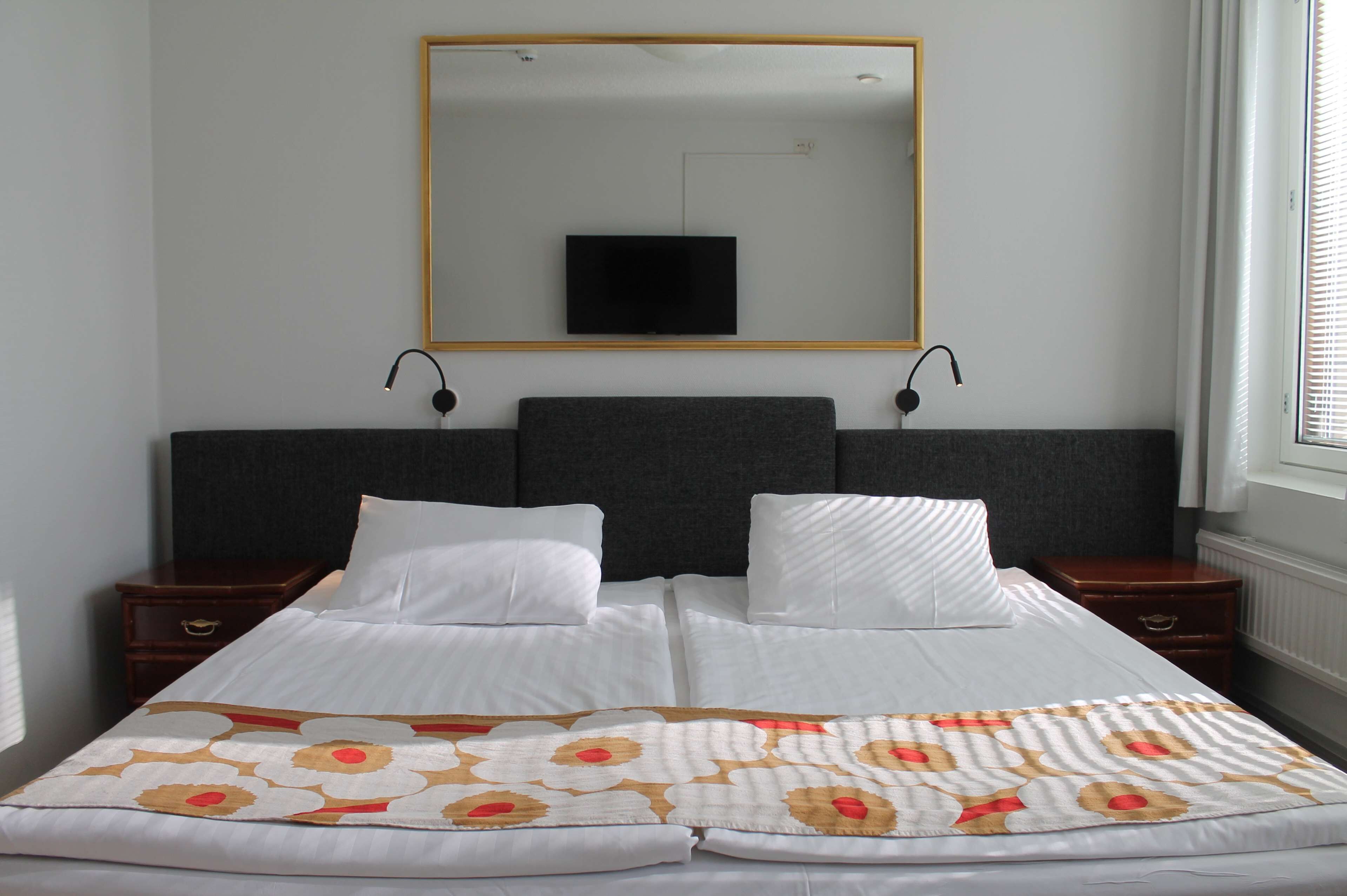 Una cama bien hecha con un edredón estampado con motivos florales, un espejo grande encima y un televisor montado en la pared.