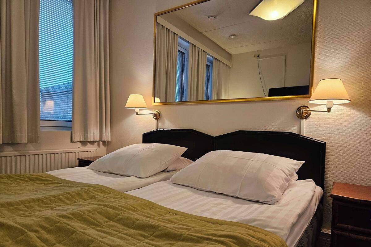 A hotel room with a double bed, two bedside lamps, a large mirror, and a window with curtains.