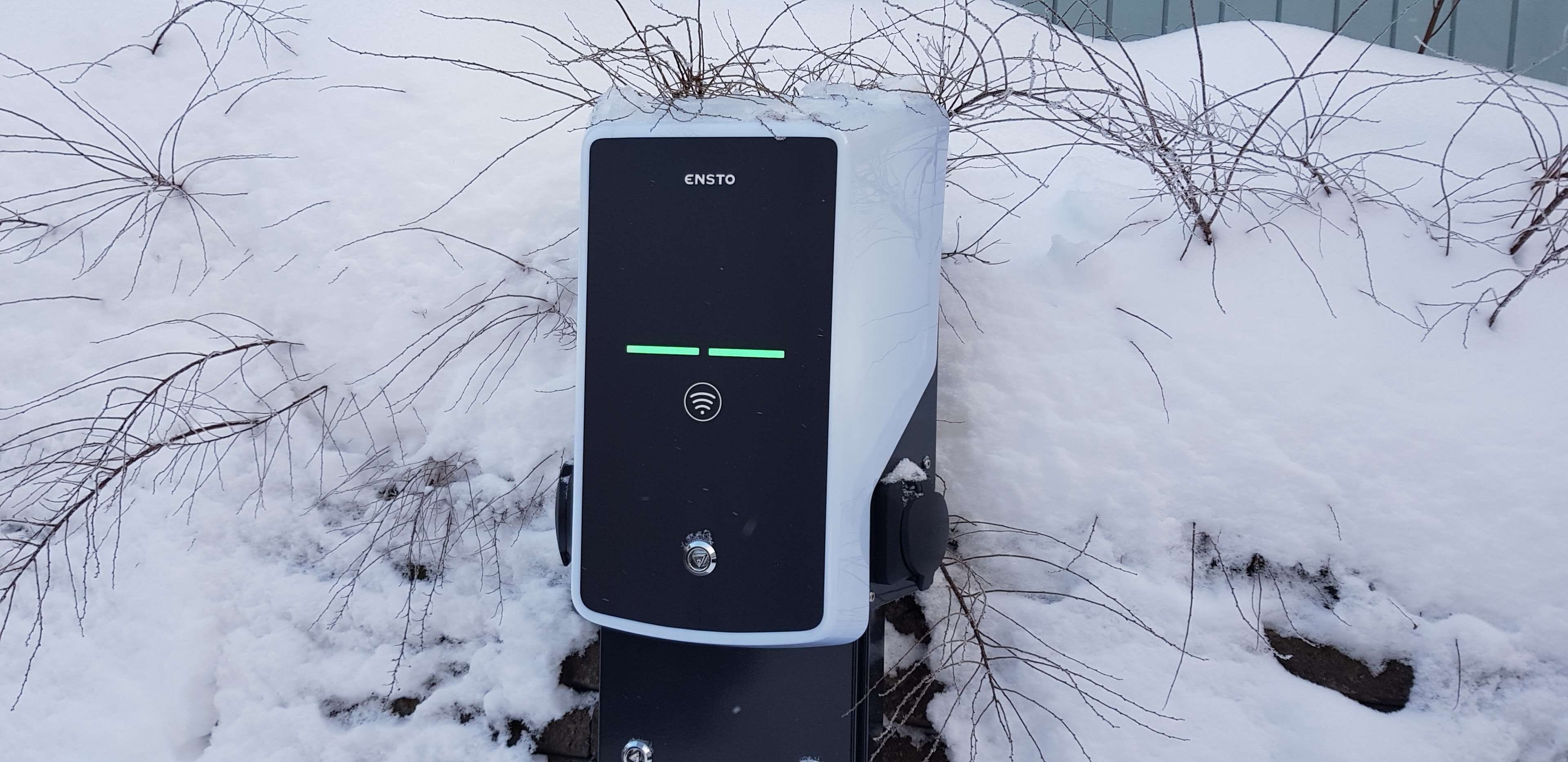 A black electric vehicle charging station with the brand ENSTO, surrounded by snow and bare branches.