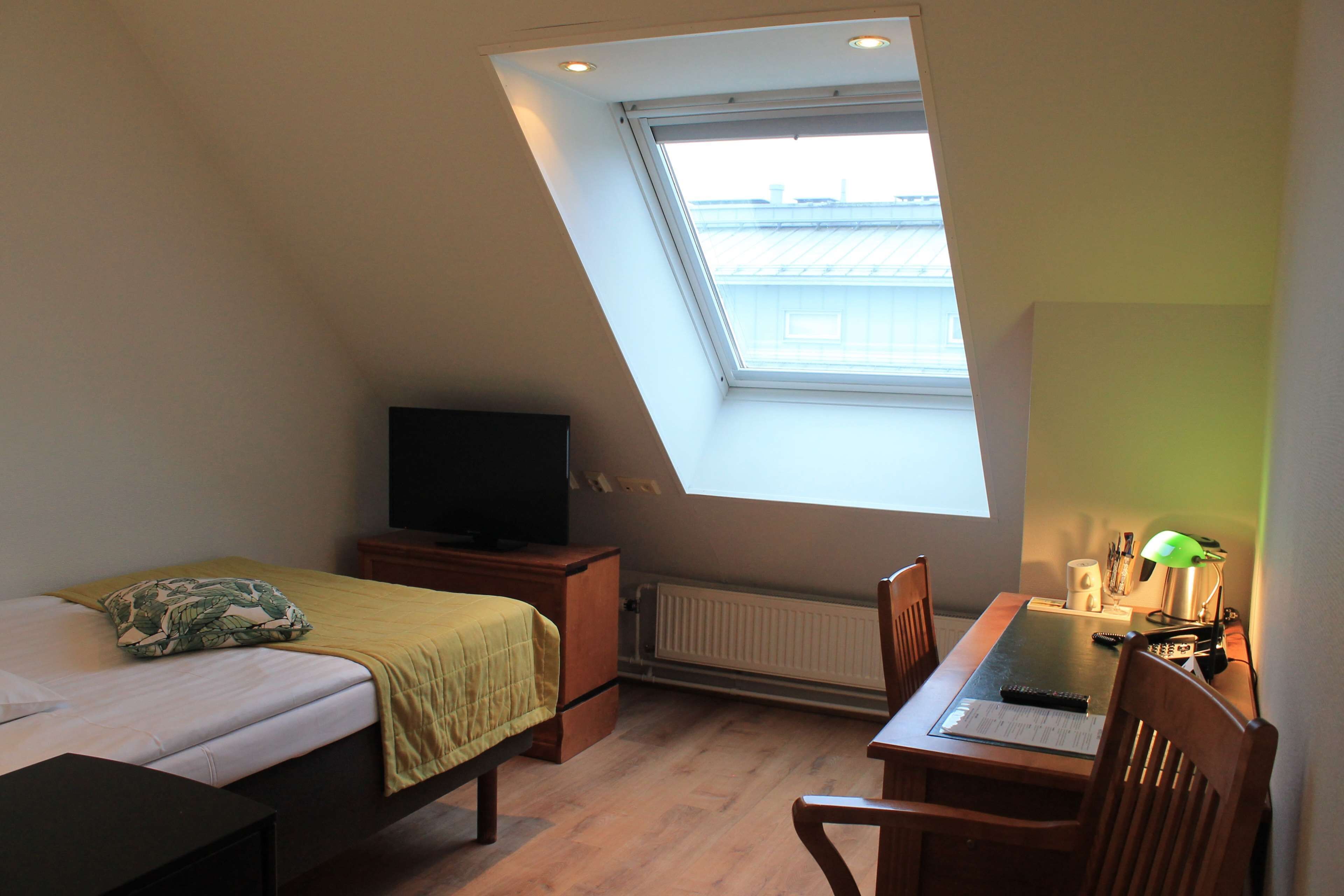 Ein Schlafzimmer mit schrägverdeckter Decke, einem Bett mit einem grünen Kissen, einem Schreibtisch mit einer Lampe und einem Fernseher auf einer Holzkommode.