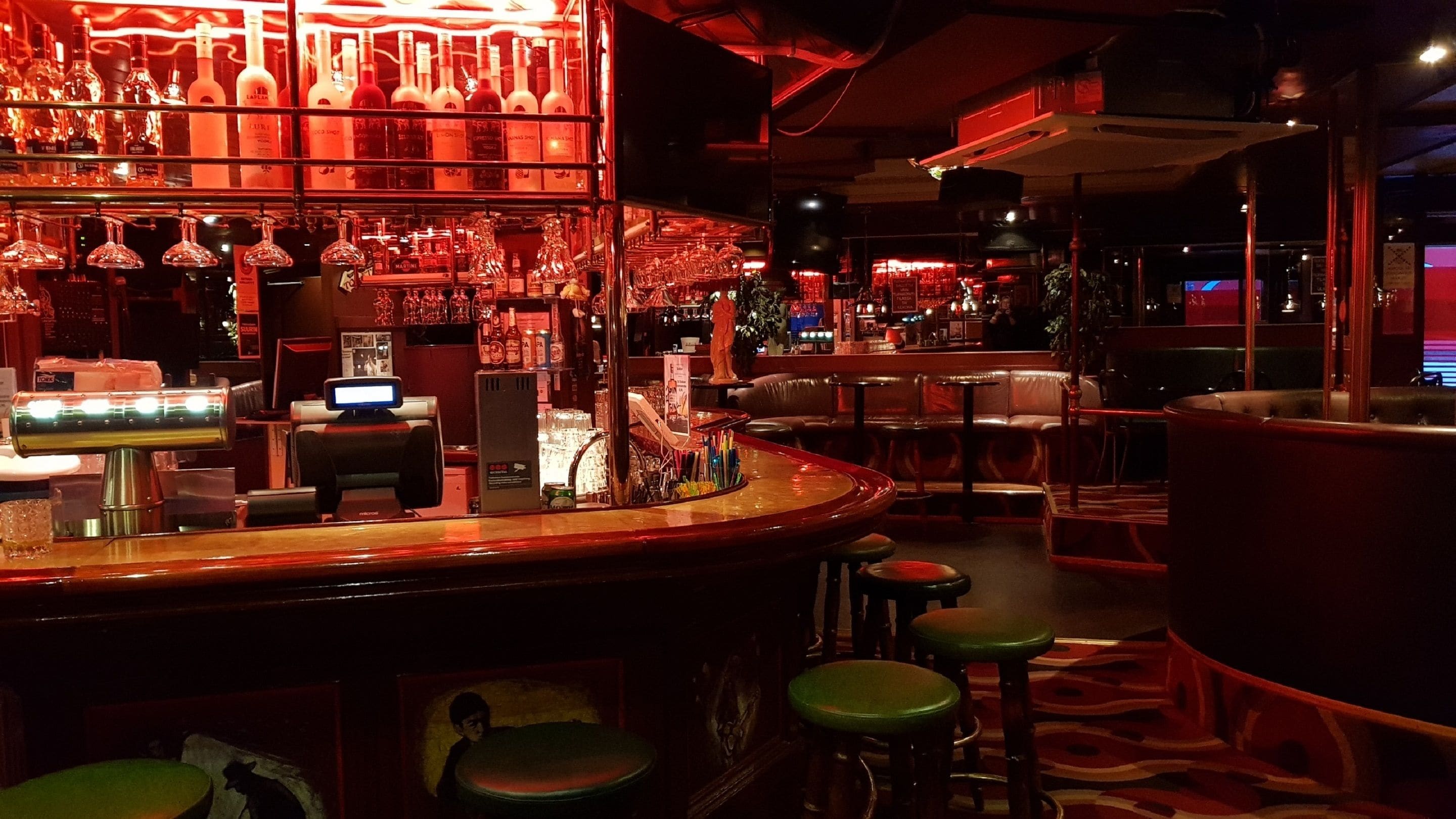 Zona de bar con estantes llenos de botellas de licores, una caja registradora y taburetes.