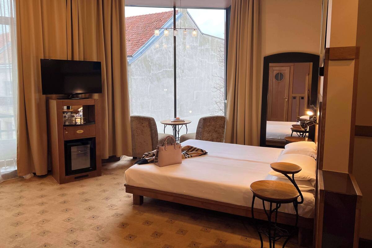 A hotel room with a large bed, a TV, a minibar, two armchairs, a small table, and a view of a building through a window.