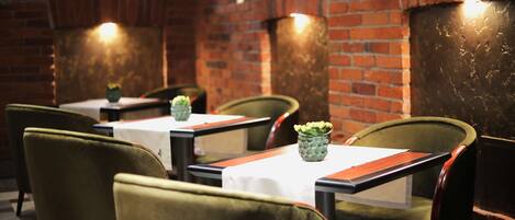 A restaurant interior with brick walls, green upholstered chairs, and small potted plants on the tables.