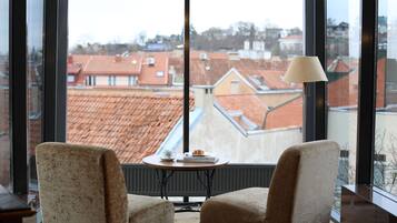 Una habitación con dos sillones, una mesita con una lámpara y vistas a tejados de tejas rojas a través de amplias ventanas.