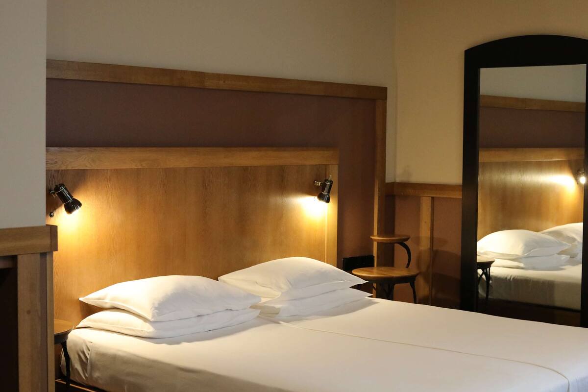 A hotel room with a wooden headboard, two bedside lamps, a round wooden table, and a large mirror.