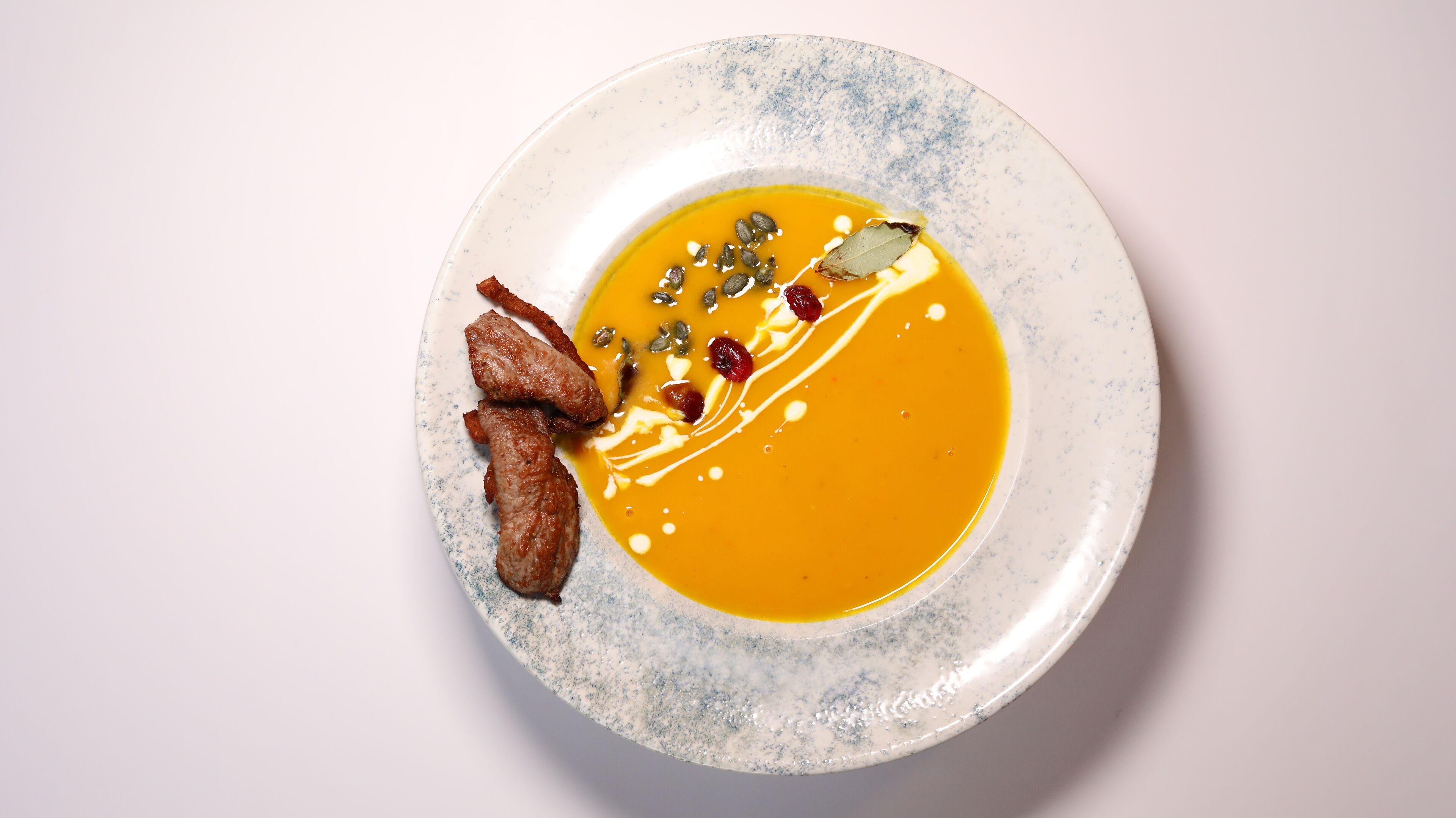Un tazón de sopa con textura cremosa, decorada con semillas y una hoja de laurel, acompañado de dos trozos de carne en un plato moteado.