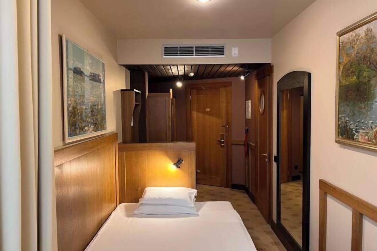 A hotel room with a bed, a wooden headboard, a wall-mounted light, a door, and a painting.