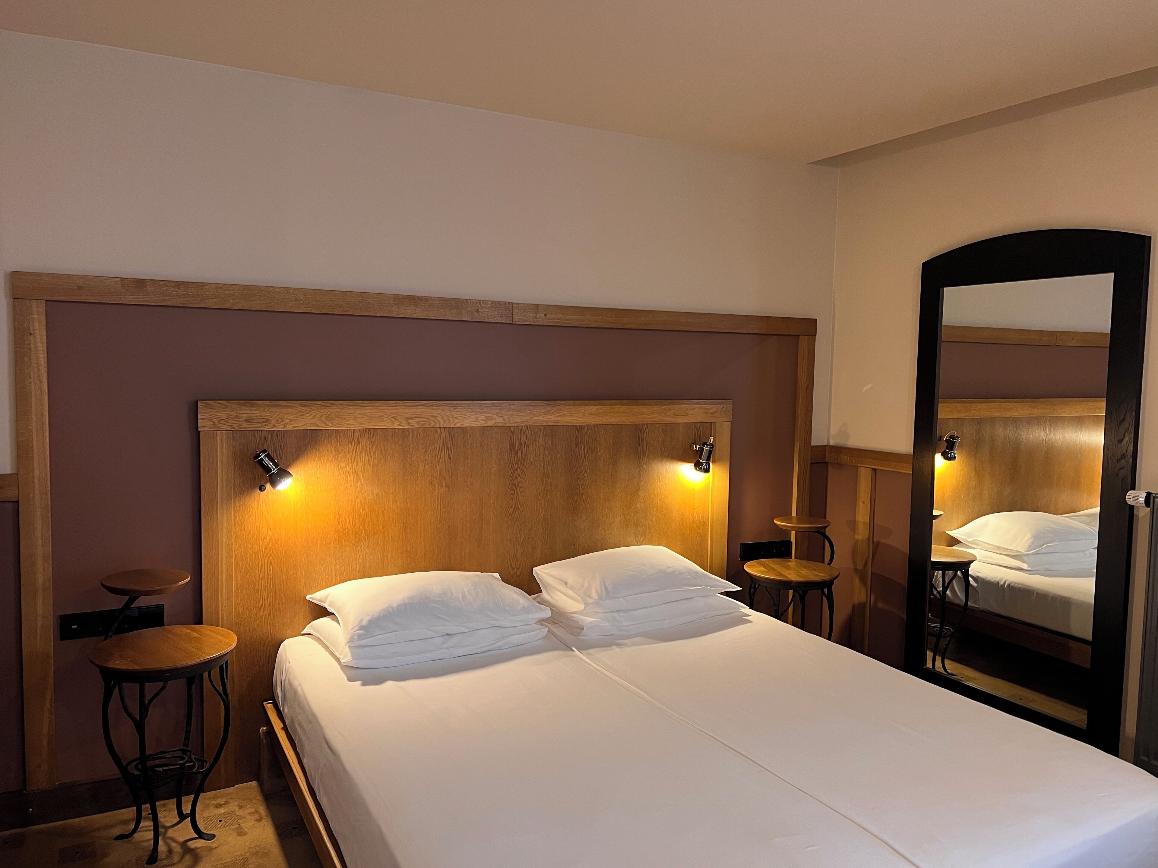 A hotel room with a wooden headboard, two bedside lamps, a bed with white linens, a round wooden side table, and a full-length mirror.