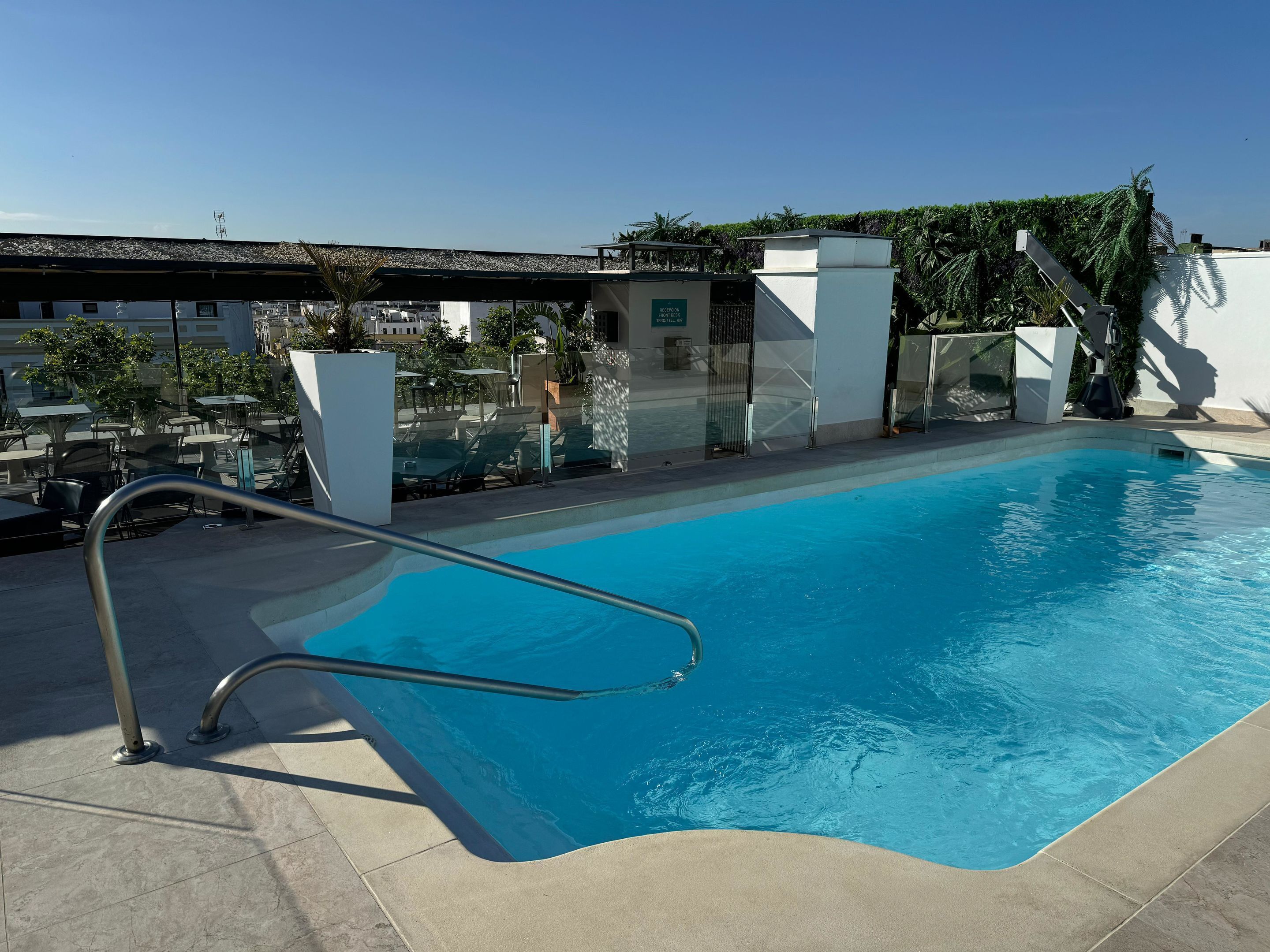 Una piscina con una escalera metálica, sillas de descanso y mesas.
