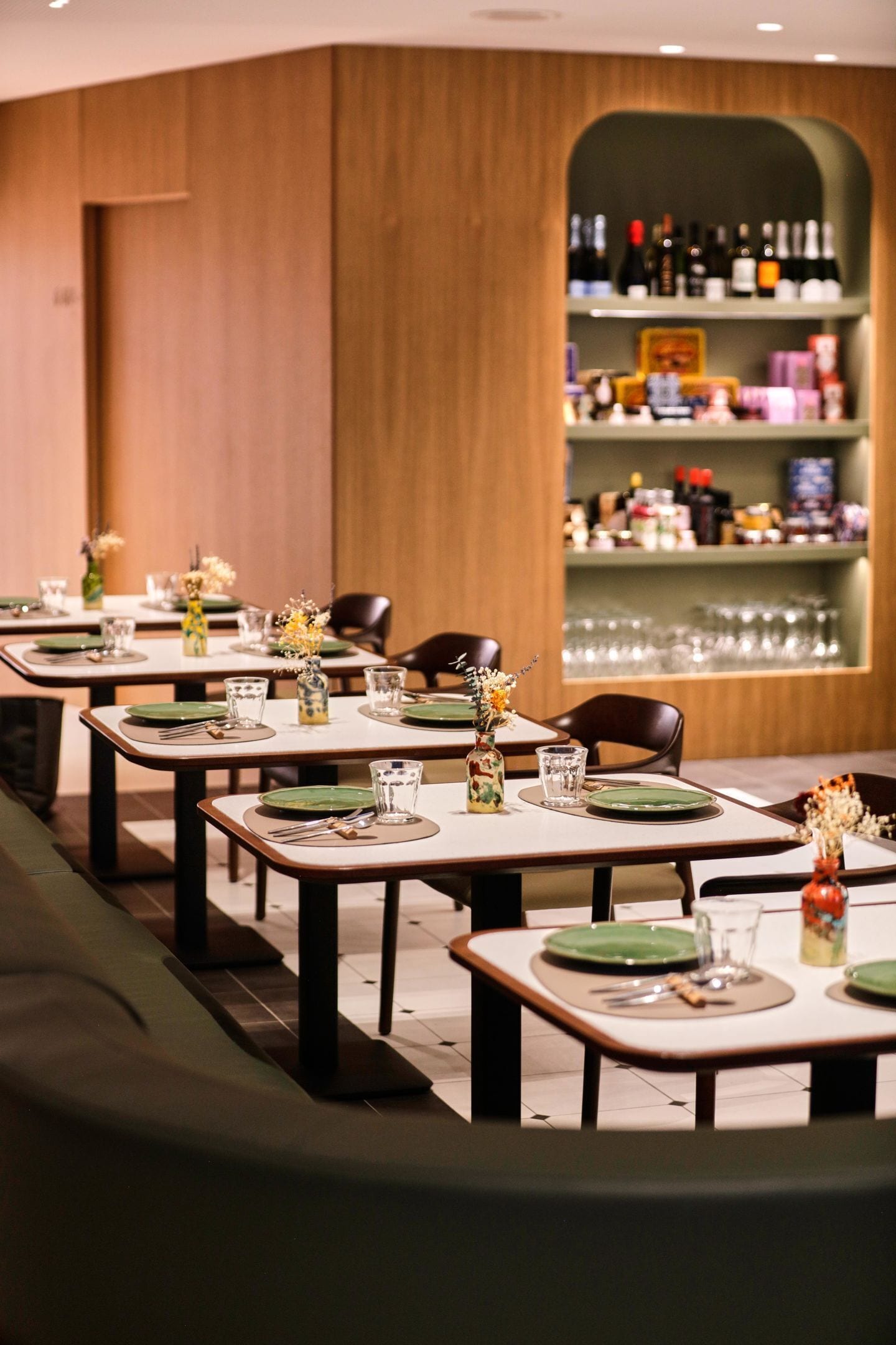 Un restaurante con varias mesas dispuestas, con platos, vasos y cubiertos verdes. Hay jarrones con flores en las mesas y una estantería para vinos al fondo.