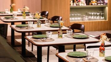 A restaurant with multiple tables set with green plates, glasses, and cutlery. There are vases with flowers on the tables, and a wine rack in the background.