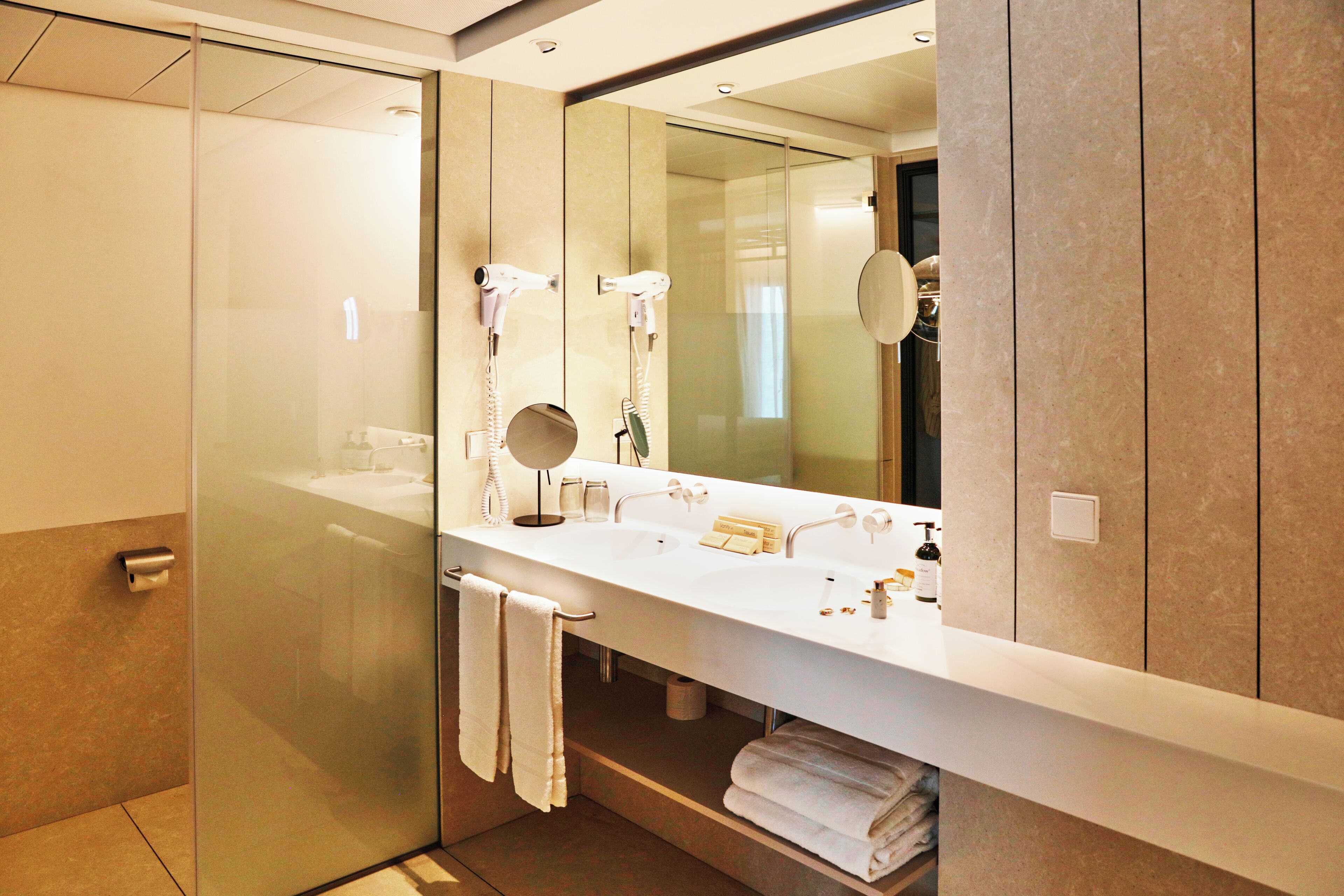 Un baño moderno con cabina de ducha de vidrio, un lavamanos blanco con espejo encima y toallas ordenadas en un estante.