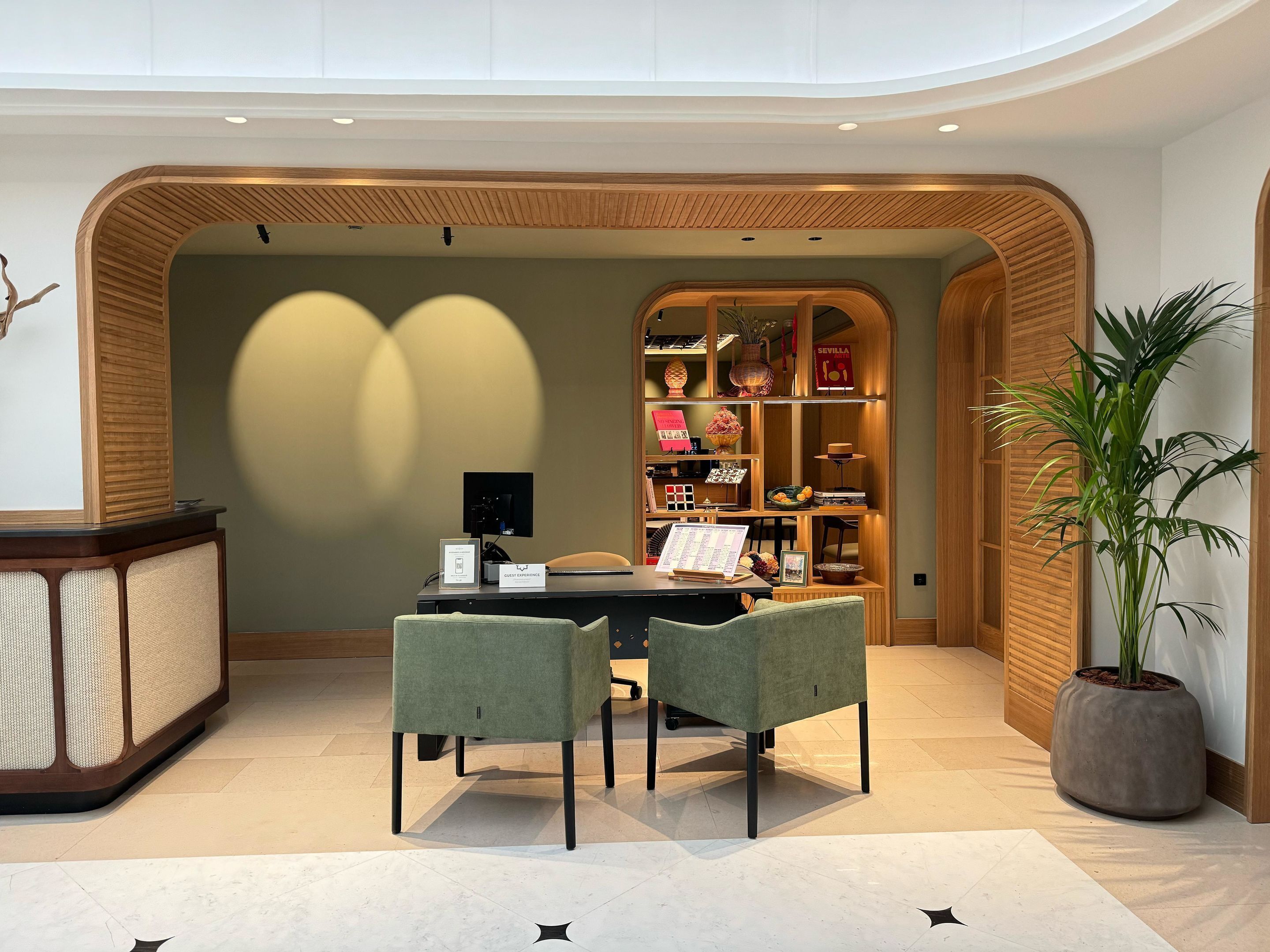 Un espacio de oficina moderno con un mostrador de recepción de madera, una computadora, dos sillas y una estantería llena de libros y objetos decorativos.