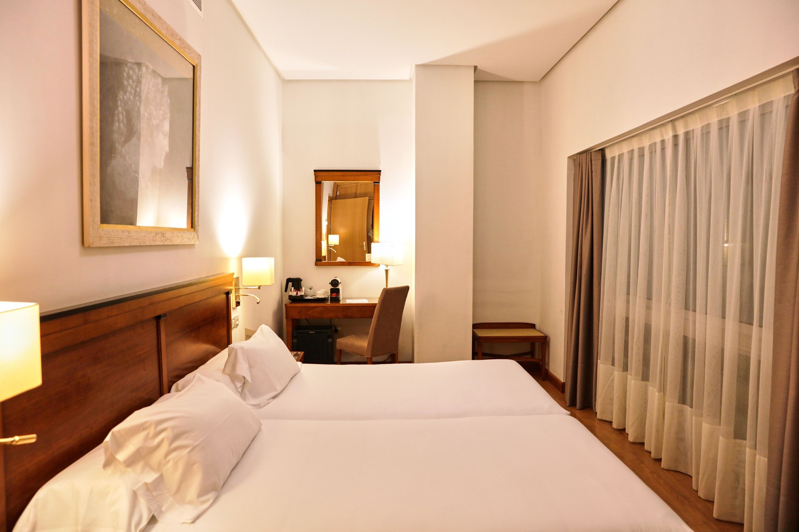 Habitació d'hotel amb un llit gran, una taula de treball, una cadira, una làmpada i una finestra amb cortines.