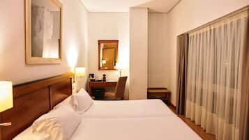 Habitació d'hotel amb un llit gran, una taula de treball, una cadira, una làmpada i una finestra amb cortines.