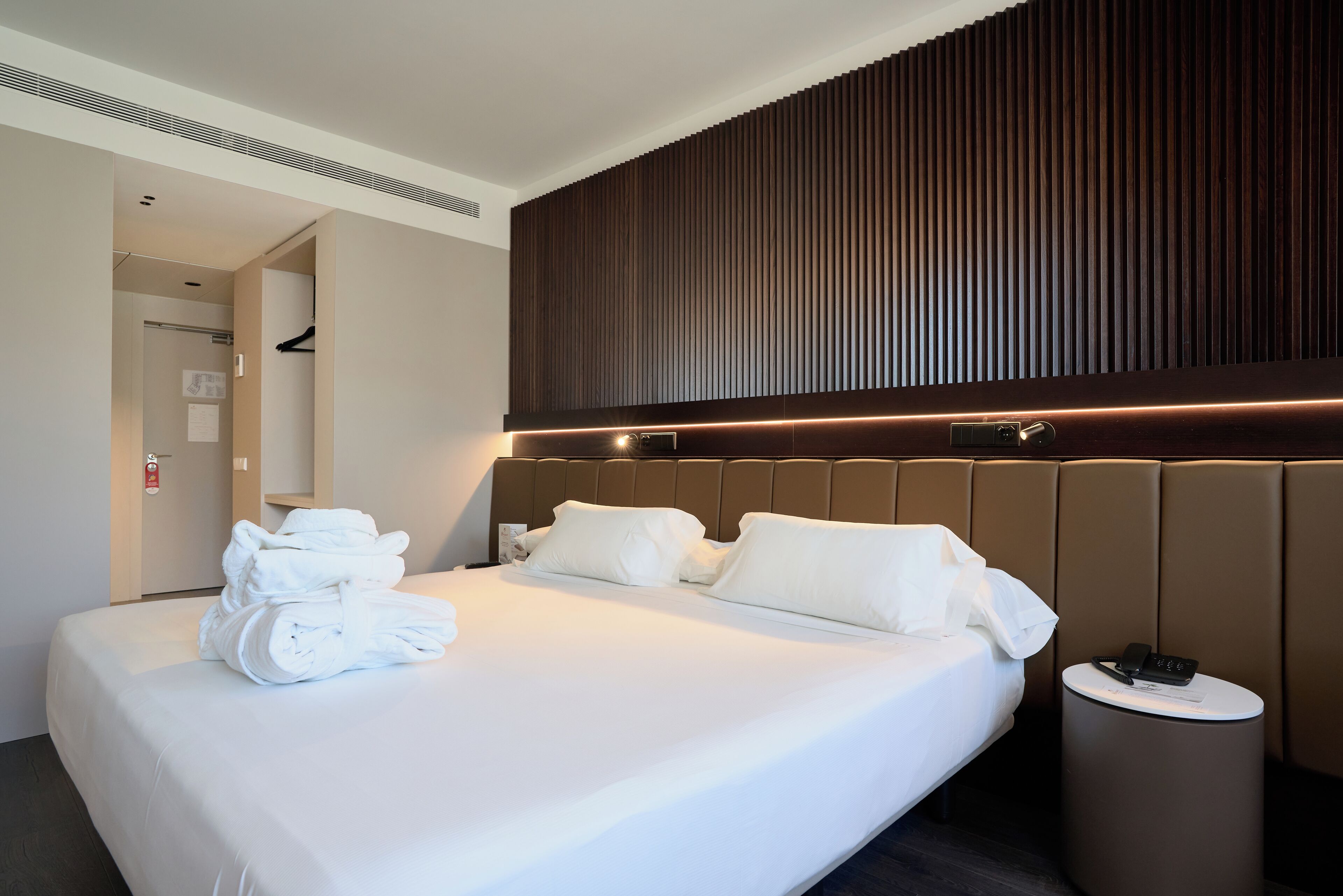 A hotel room with a large bed, a headboard with vertical lines, a bedside table with a phone, and a wall-mounted light.