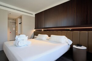 A hotel room with a large bed, a headboard with vertical lines, a bedside table with a phone, and a wall-mounted light.