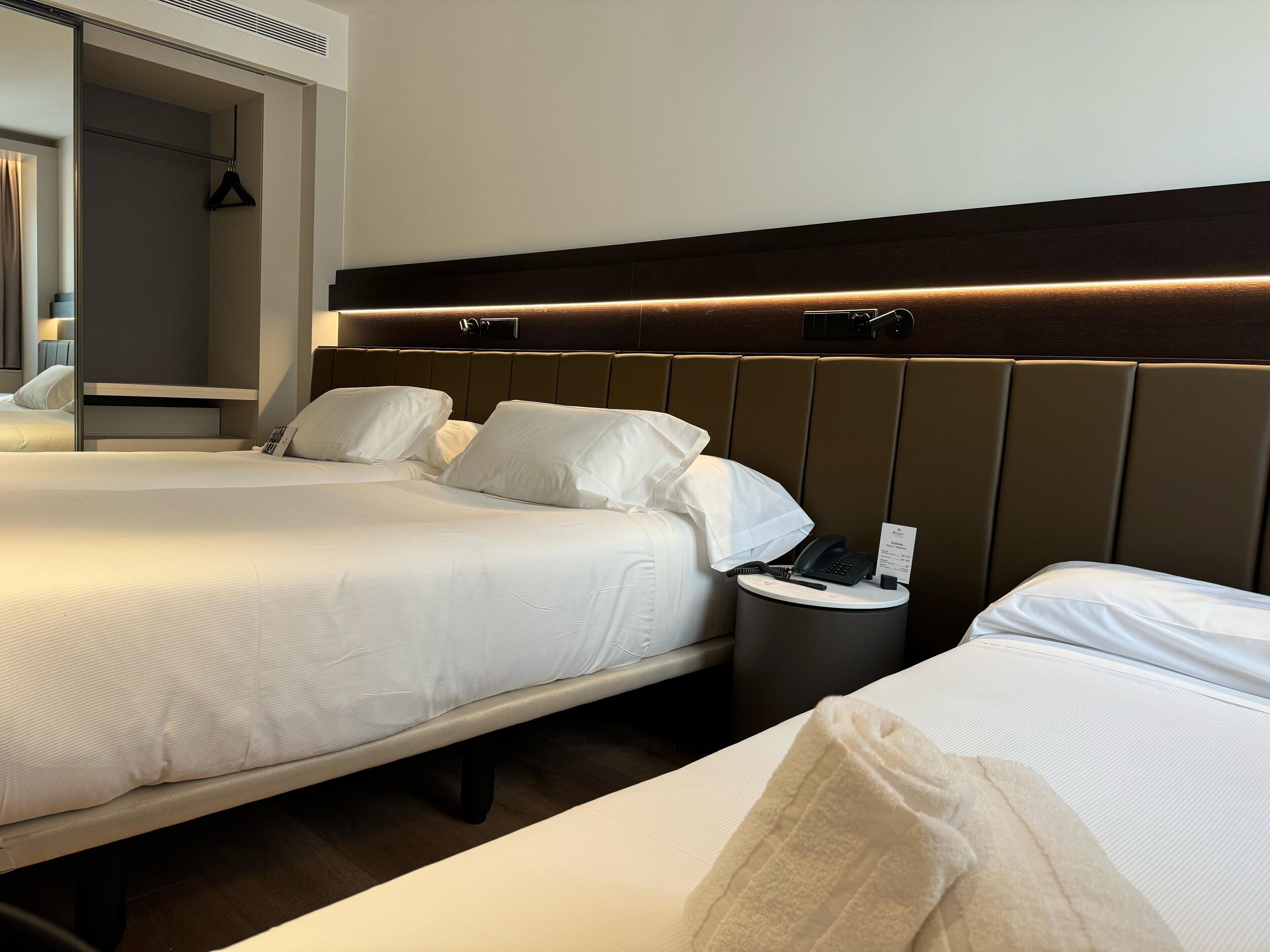 A hotel room with two beds, a bedside table with a phone and a sign, and a mirror with a hanger.