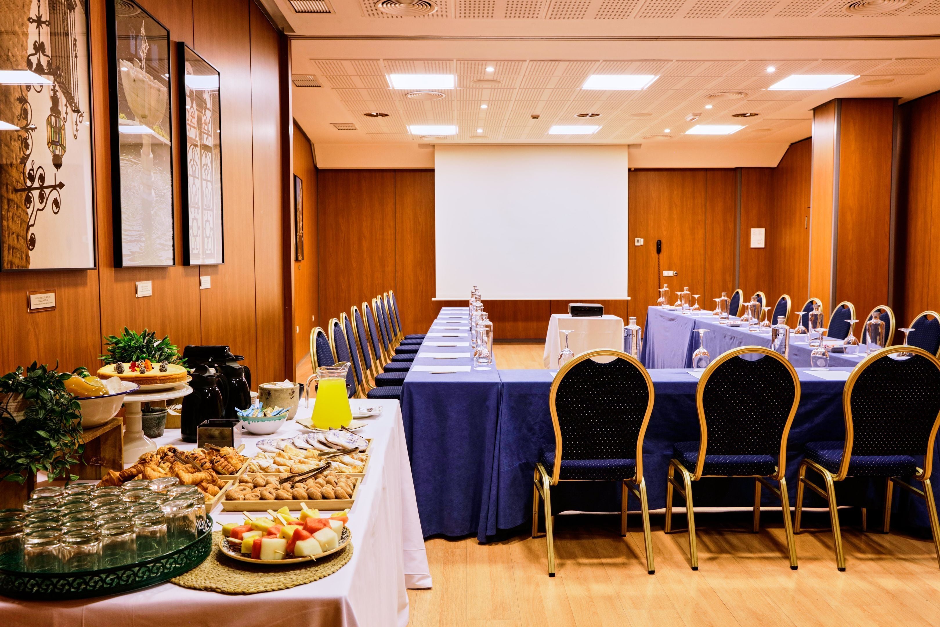 A conference room with a long table set for a meeting, equipped with chairs, a projector screen, and a buffet with various food items.