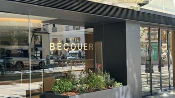 Uma entrada de hotel moderna com fachada de vidro, um letreiro com a inscrição "HOTEL BÉCQUER SEVILLA" e um vaso com plantas em frente.