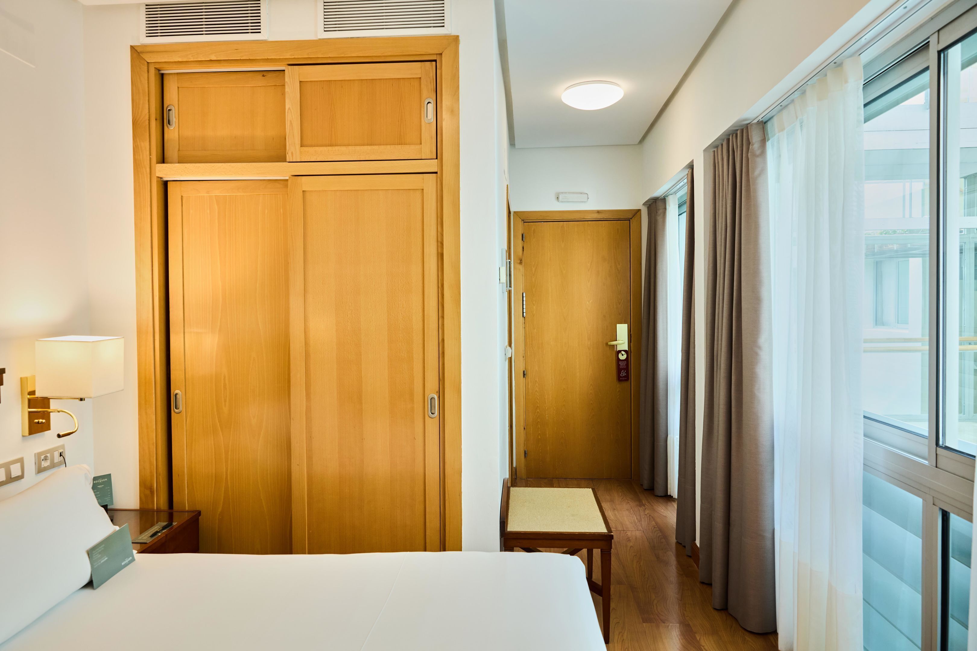 Ein Hotelzimmer mit Bett, Holzschrank, Nachttisch, Lampe und Blick durch ein Fenster mit Vorhängen.