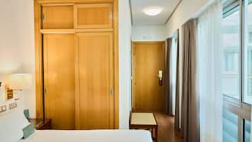 Ein Hotelzimmer mit Bett, Holzschrank, Nachttisch, Lampe und Blick durch ein Fenster mit Vorhängen.