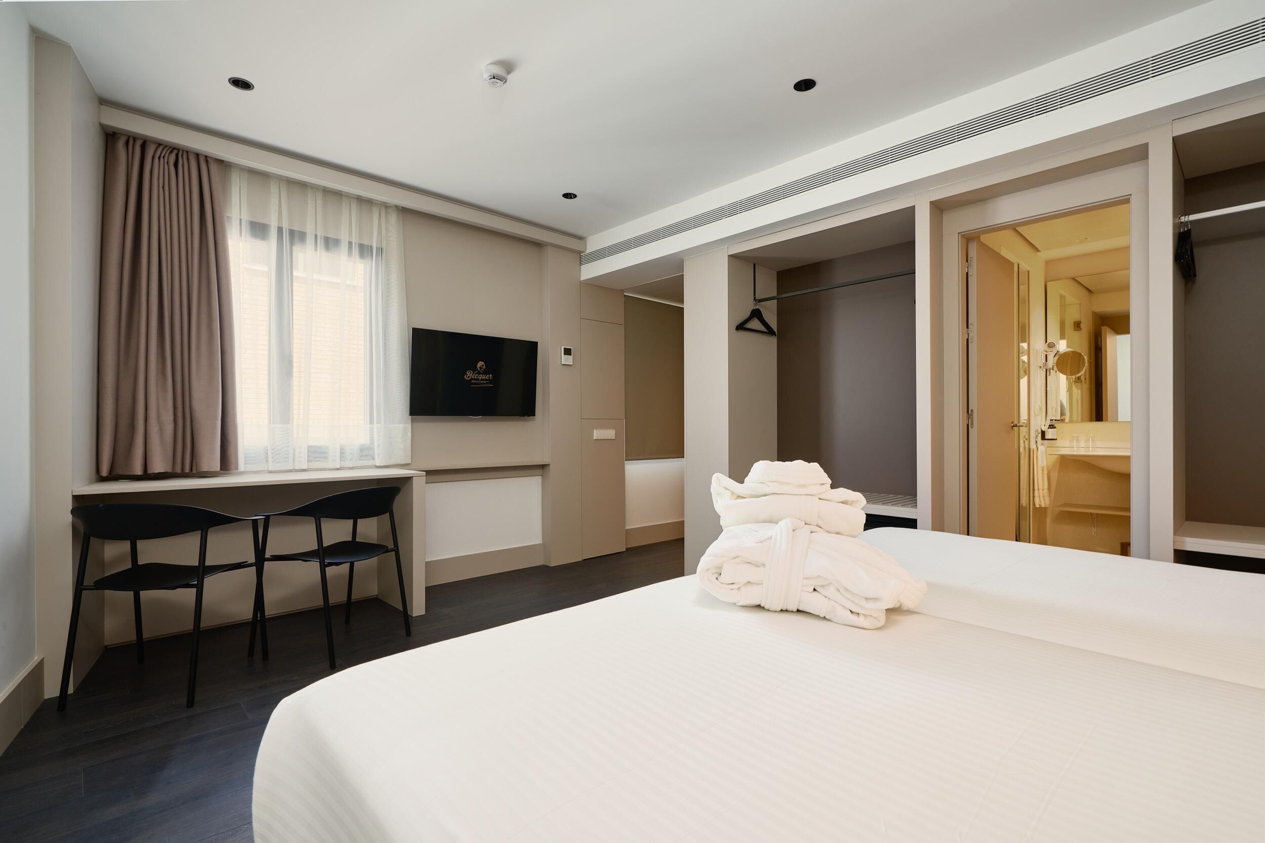 Habitación de hotel con cama, escritorio con dos sillas, televisor y armario con espejo.
