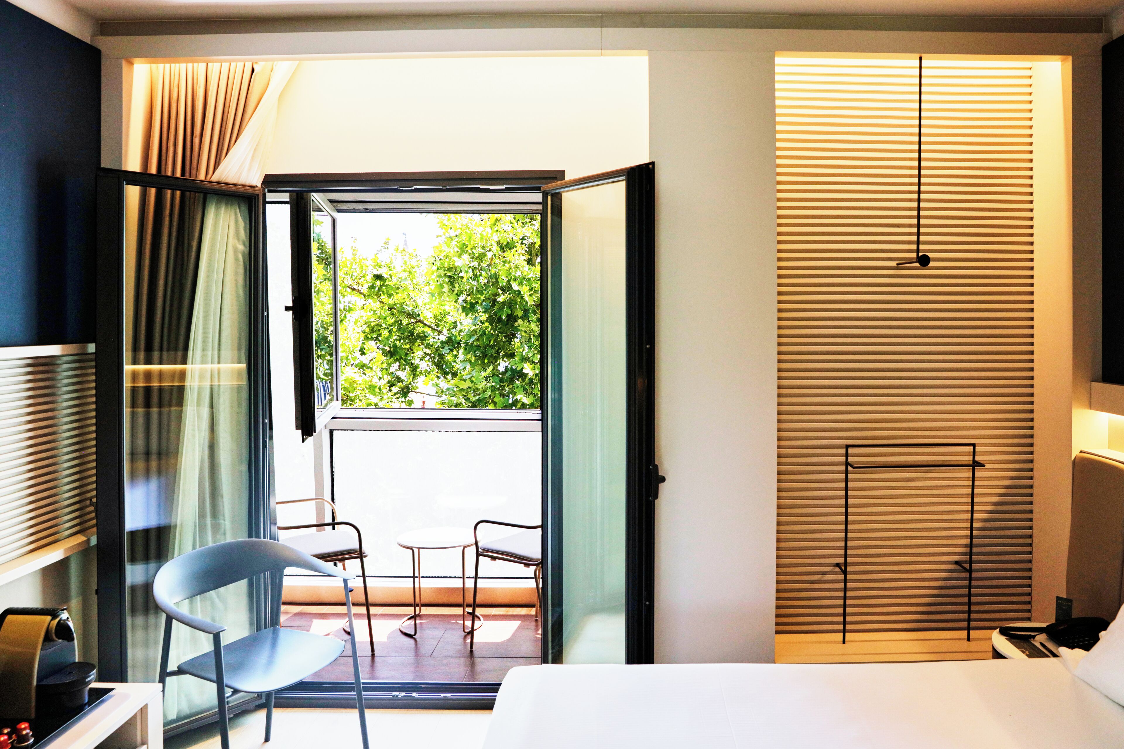 A hotel room with a bed, a desk with a telephone, a chair, and a view of greenery through an open door.