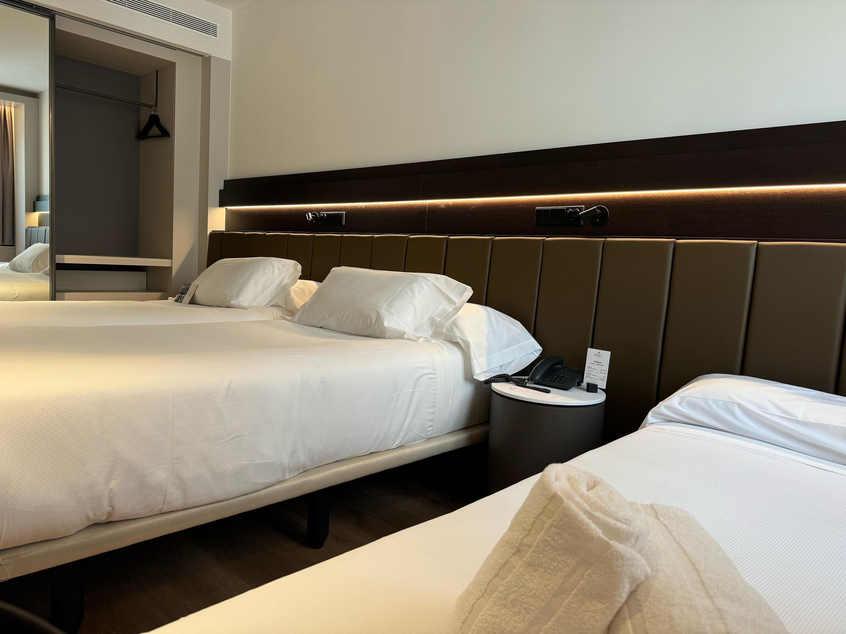 A hotel room with two beds, a bedside table with a phone and a sign, and a mirror.