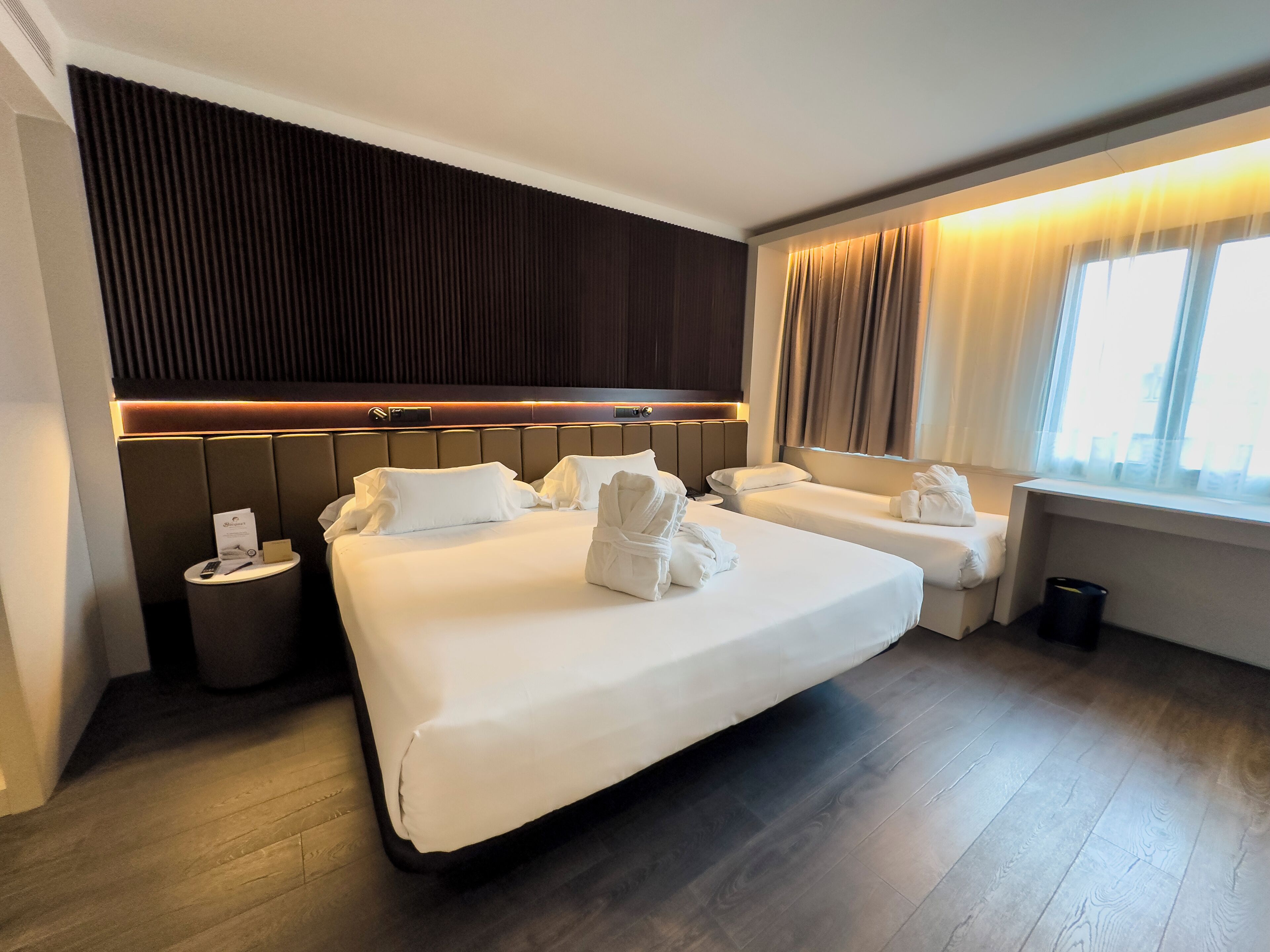 A hotel room with a large bed, a smaller bed, a bedside table, and a window with curtains.