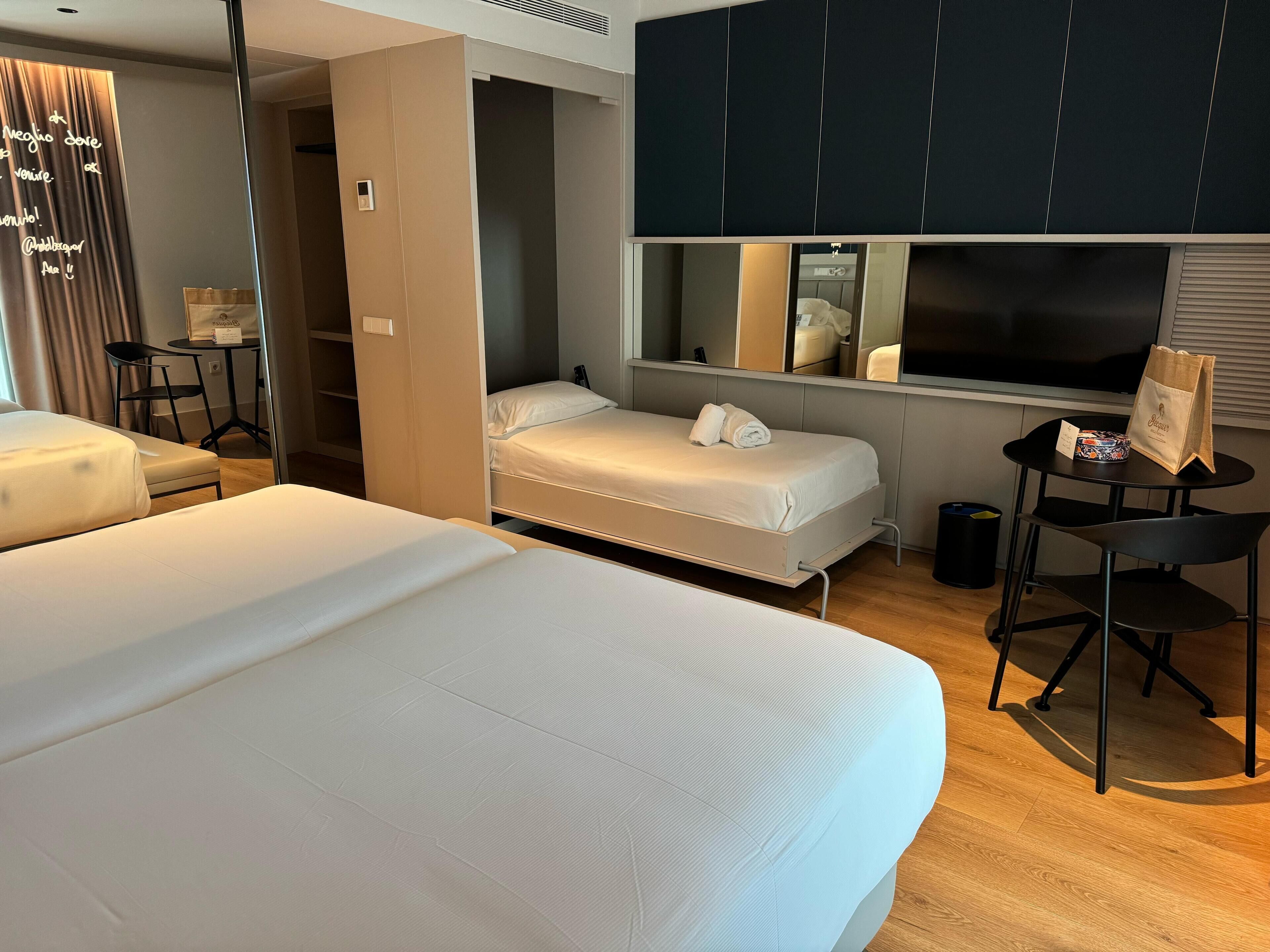 Habitación de hotel con dos camas, un escritorio, una silla, una mesita, un bote de basura y un espejo.