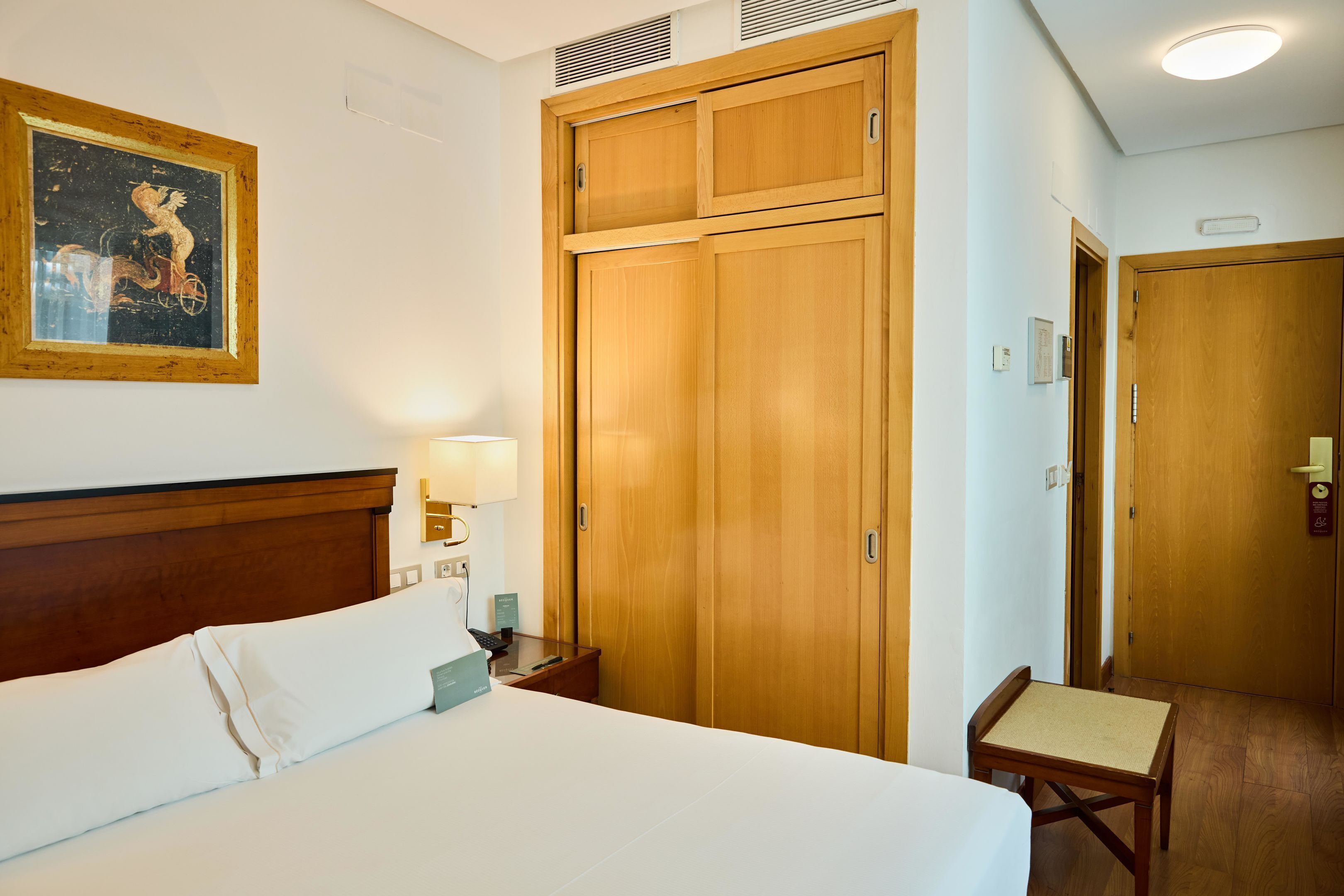 Habitació d'hotel amb un llit, un armari de fusta, una taula de treball amb una làmpada i un quadre al mur.