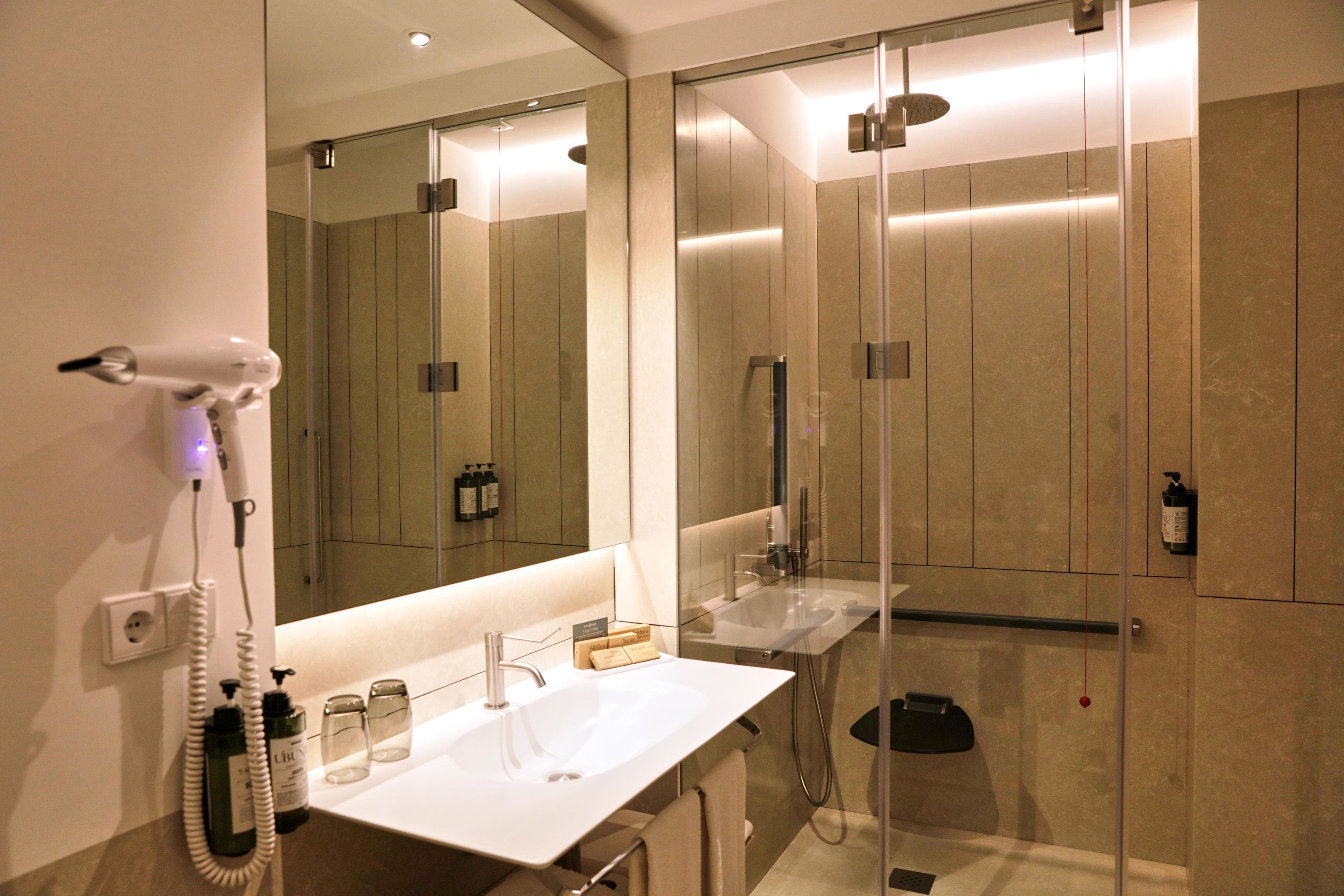 Un baño moderno con un espejo grande, un lavamanos blanco, una cabina de ducha de vidrio y un secador de pelo en la pared.
