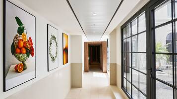 A hallway with framed artwork on the walls, a tiled floor, and large windows on the right.