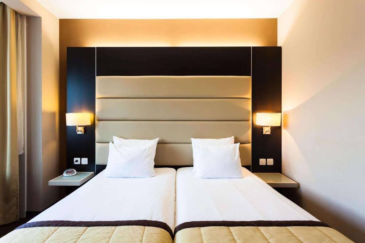 Habitación de hotel con dos camas, un cabecero grande, lámparas de noche y una mesita de noche con un teléfono.