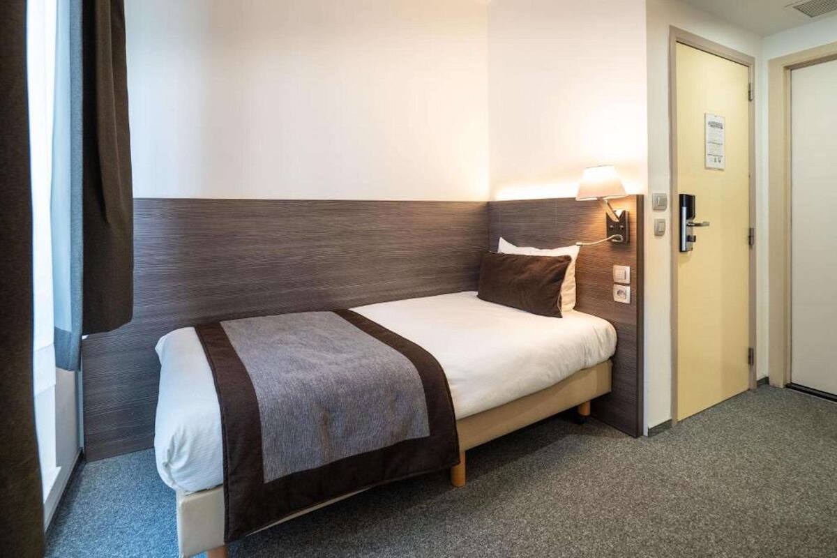 Habitación de hotel con cama individual, mesita de noche con lámpara, puerta con cerradura e interruptor de luz en la pared.