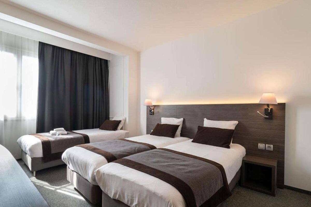 Habitación de hotel con dos camas, un ventanal con cortinas, una lámpara de pared y una mesita de noche pequeña.