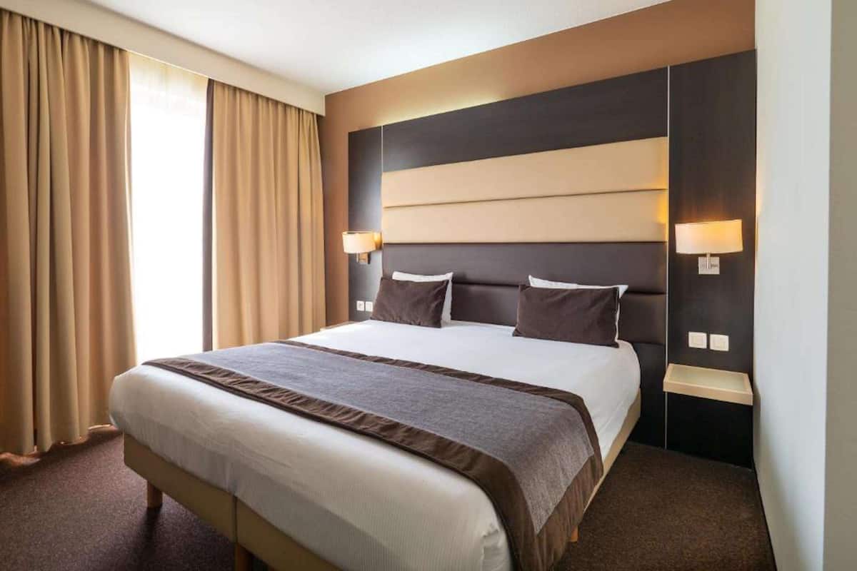 Habitación de hotel con una cama grande, dos mesitas de noche, cabecero y una ventana con cortinas.