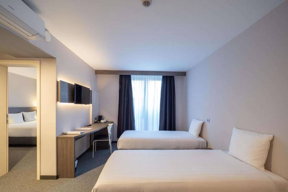 Habitación de hotel con dos camas, un escritorio, una silla, un televisor de pantalla plana y una ventana con cortinas.