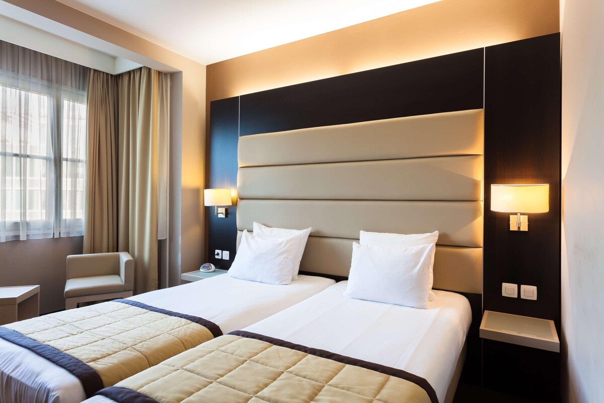 A hotel room with two beds, a nightstand, a chair, and a window with curtains.