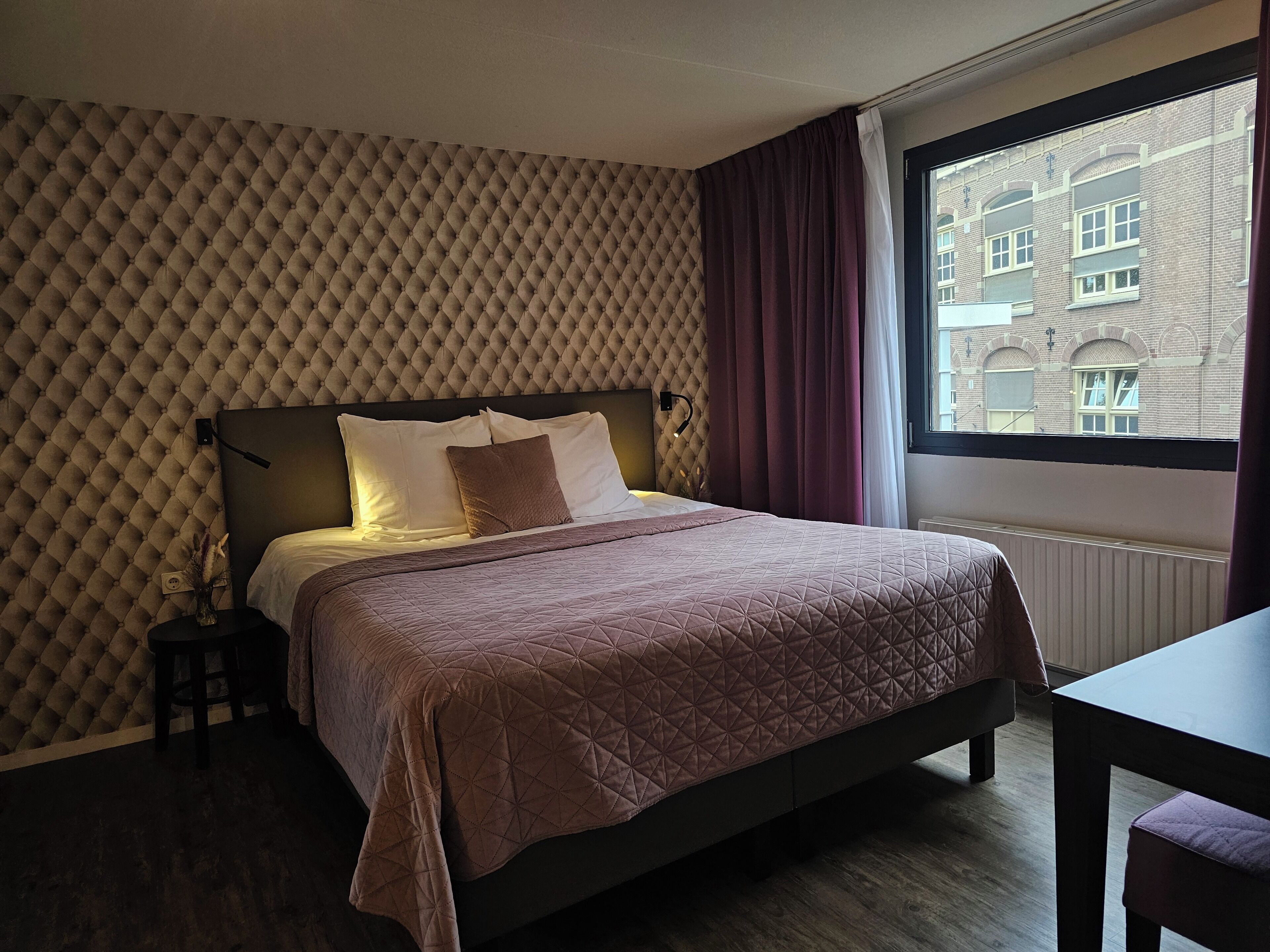 Un lit bien fait, recouvert d’une courtepointe, une tête de lit à motifs capitonnés, une table de chevet avec une lampe et une fenêtre donnant sur des immeubles.