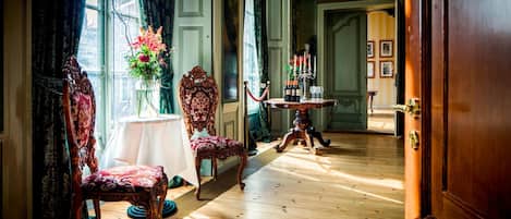 Una habitación con suelo de madera, una mesa con un florero y dos sillas ornamentadas.