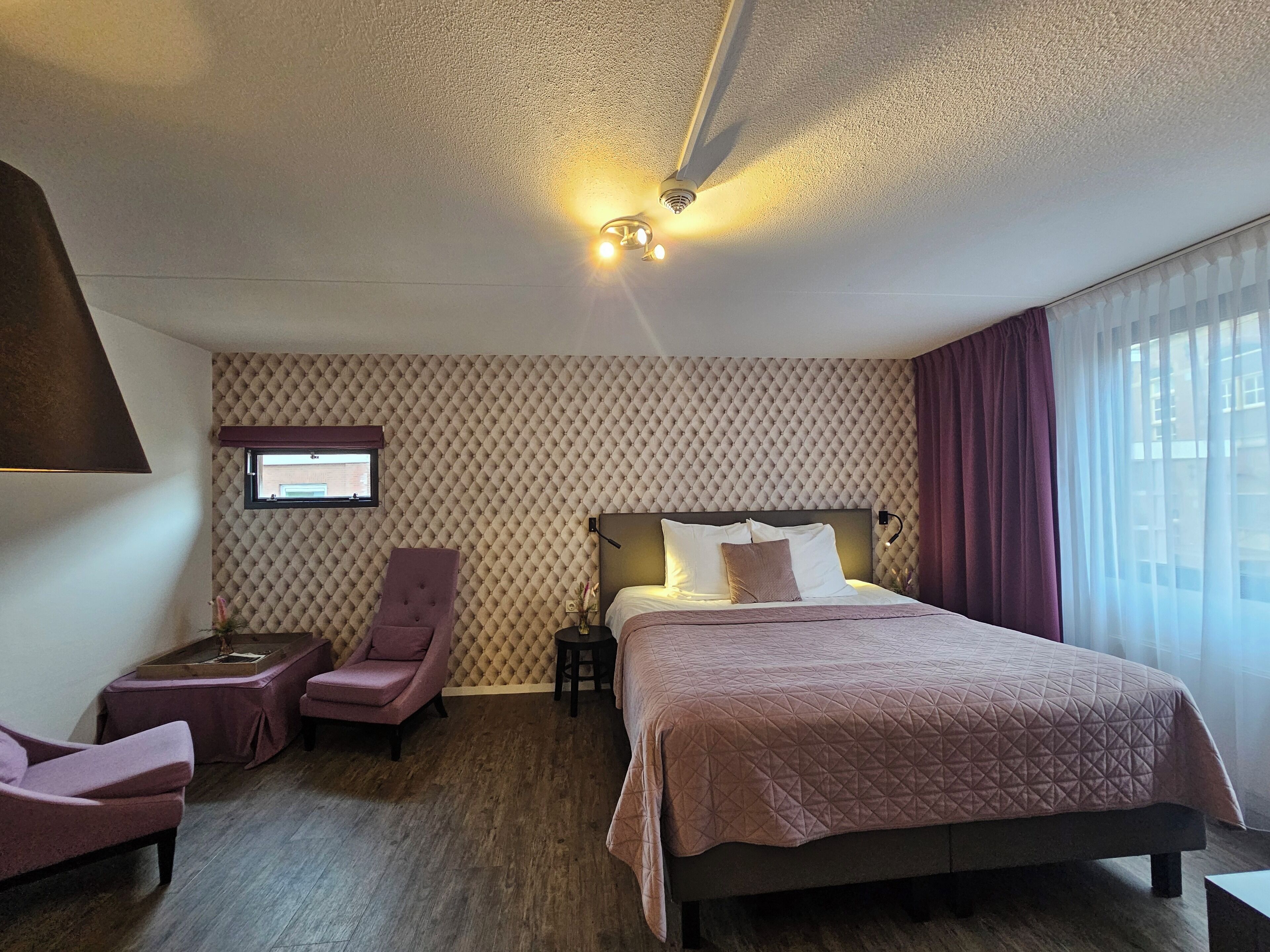 A hotel room with a bed, a chair, a small table, a TV, and a window with curtains.