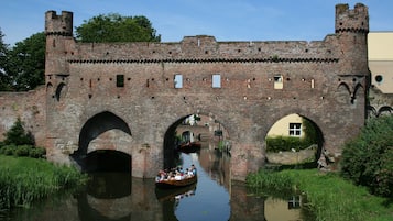 Un puente de ladrillo histórico con tres arcos sobre un canal, una embarcación con pasajeros y un edificio similar a un castillo al fondo.
