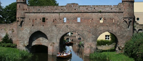 Un puente de ladrillo histórico con tres arcos sobre un canal, una embarcación con pasajeros y un edificio similar a un castillo al fondo.